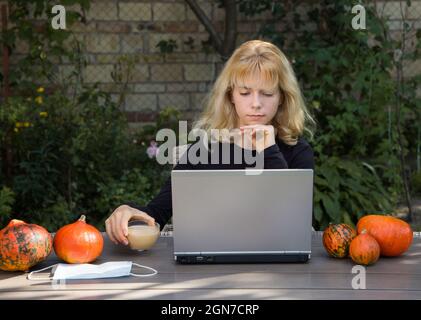 Porträt einer jungen Frau, die im Garten am Tisch sitzt, am Laptop arbeitet und Cappuccino trinkt. Halloween Dekorationen aus orangefarbenen Kürbissen. Arbeiten, studieren bei Stockfoto