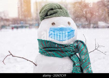 Lustiger Schneemann in schützender medizinischer Maske auf weißem Hintergrund. Weihnachten Hintergrund während Coronavirus. Schnee Figur mit grün gestrickt Schal und Hut.. Stockfoto