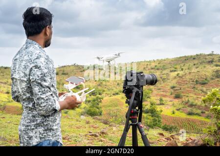Fokus auf Drohne, junger Videofilmer, der Videos filmt, indem er die Drohne mithilfe der Fernbedienung steuert - Konzept der professionellen Drohnenfotografie und -Antenne Stockfoto