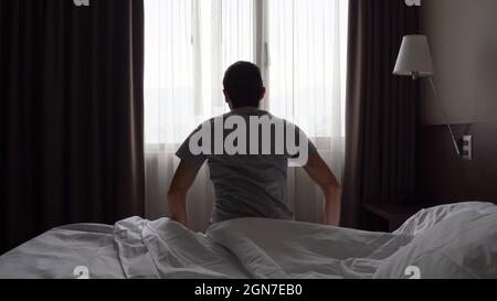 Silhouette eines einsamen Mannes, der im dunklen Zimmer am Schlafzimmerfenster auf dem Bett sitzt. Depressiv, am frühen Morgen aufwachen, faul, müde Konzepte Stockfoto