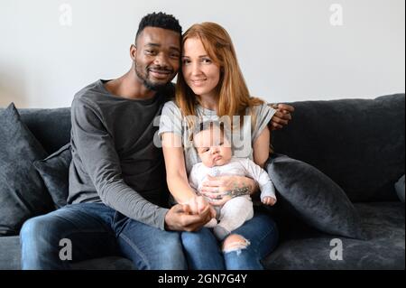 Porträt einer glücklichen gemischten Familie aus drei afroamerikanischen Ehemännern, kaukasischer Frau und einer niedlichen Birazialtochter, die drinnen auf der Couch sitzt und die Kamera anschaut Stockfoto