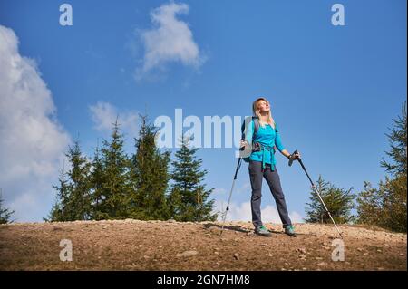 Reisende Frau mit Rucksack, die auf einer Steinstraße steht und sich mit geschlossenen Augen unter Sonnenstrahlen sonnt, während sie im Freien wandert. Touristische Sommerwanderung in der Natur. Stockfoto