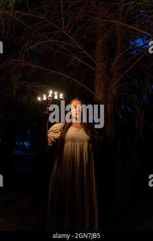 Eine junge Frau im dunklen Wald mit einem Kerzenleuchter. Trägt ein altes langes weißes Kleid. Stockfoto