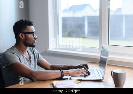 Ein intelligenter afroamerikanischer Mann, der eine Brille und ein legeres T-Shirt trägt und einen Laptop für ferngesteuerte Arbeit am Schreibtisch zu Hause verwendet. Ein junger schwarzer Student, der online studiert, aus der Ferne lernt, Seitenansicht Stockfoto
