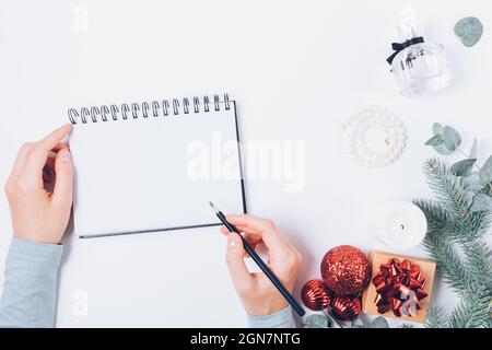 Weibliche Hände schreiben in leerem Notizbuch neben Weihnachtsdekorationen, Geschenkbox, Tannenzweigen und femininen Accessoires auf weißem Tisch, Draufsicht. Stockfoto