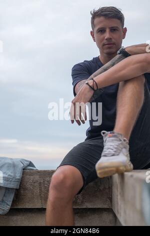 Porträt eines jungen hübschen kaukasischen Mannes in lässigem Tragen und Sneakers. Sitzen und auf die Kamera mit Kopierplatz schauen Stockfoto