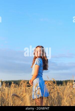 Glückliches junges Mädchen lächelt auf Weizenfeld und berührt Weizenohren mit der Hand. Schöner Teenager, der die Natur bei warmem Sonnenschein in einem Weizenfeld genießt Stockfoto