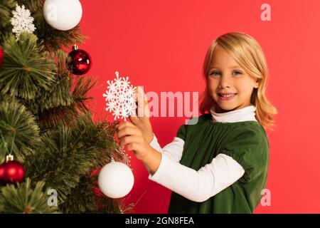 Glückliches Kind hält dekorative Schneeflocke in der Nähe weihnachtsbaum isoliert auf rot Stockfoto