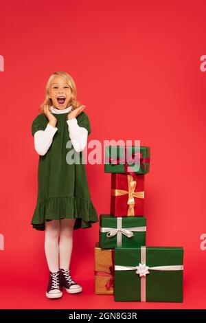 Erstauntes Kind in grünem Kleid und Gummischuhen, die Hände klatschen, neben einem Stapel von Geschenken auf rot Stockfoto
