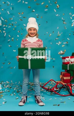 Fröhliches Kind im warmen Hut mit großer Geschenkbox neben Konfetti und Schlitten mit Geschenken auf blau Stockfoto