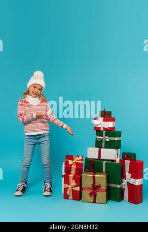 Lächelndes Mädchen in Strickmütze, Pullover und Jeans, das auf den Stapel von Geschenken auf Blau zeigt Stockfoto