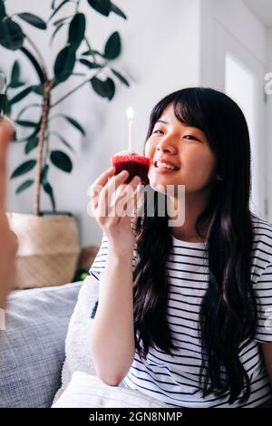 Lächelnde junge Frau, die zu Hause Cupcake mit Kerze hält Stockfoto