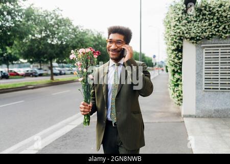 Lächelnder Geschäftsmann hält Blumenstrauß, während er auf dem Handy auf der Straße telefoniert Stockfoto