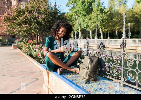 Lächelnde weibliche Touristin, die Mobiltelefon benutzt, während sie auf der Bank auf der Plaza De Espana, Sevilla, Spanien sitzt Stockfoto