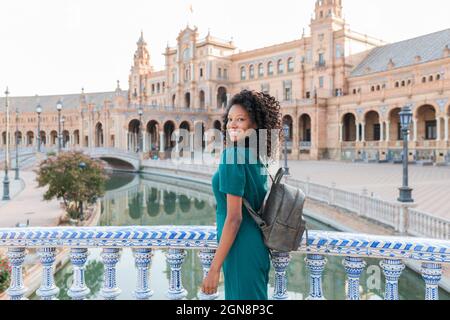 Lächelnde junge Frau mit lockigen Haaren, die am Geländer auf der Plaza De Espana, Sevilla, Spanien, steht Stockfoto