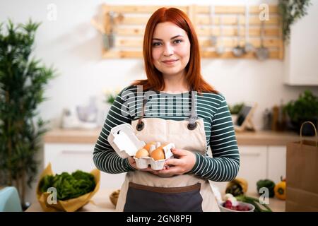 Lächelnde Rothaarige Frau hält Eierkarton in der Küche Stockfoto