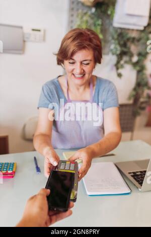 Lächelnde, reife Floristin mit Kreditkartenleser, während der Kunde beim Checkout kontaktlos über das Mobiltelefon bezahlt Stockfoto