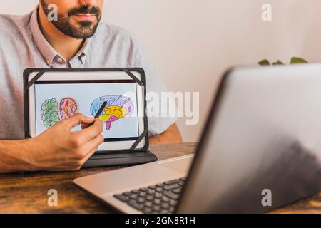 Männlicher psychischer Gesundheitsexperte, der während der Online-Therapie ein Gehirndiagramm auf einer digitalen Tablette zeigt Stockfoto