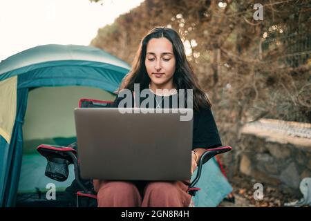 Lächelnde junge Frau mit Laptop, während sie auf einem Stuhl im Wald sitzt Stockfoto