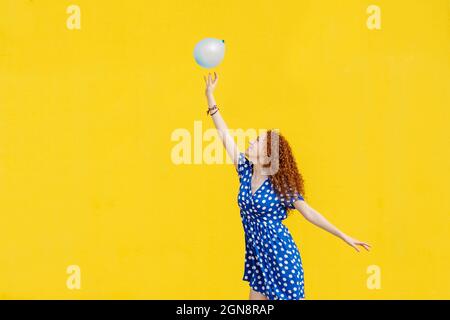 Sorglose Rotschopf-Frau, die mit einem Ballon vor einer gelben Wand spielt Stockfoto