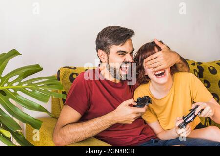 Fröhlicher Mann, der die Augen seiner Freundin bedeckt, während er zu Hause ein Videospiel spielt Stockfoto