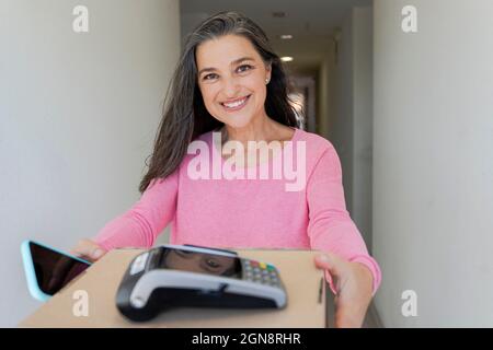 Lächelnde Frau mit Mobiltelefon, die das Paket auf dem Flur entgegenbekommt Stockfoto