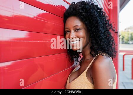 Lächelnde junge Frau mit lockigen Haaren, die auf einem roten Verschluss gelehnt ist Stockfoto