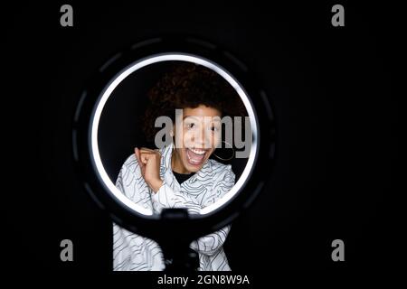 Glückliche trendige Frau, die durch Ringlicht vor schwarzem Hintergrund schaut Stockfoto