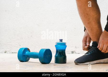 Mann bindet seine Schuhe neben Hantel und Flasche mit blauem Wasser, vor dem Training. Übung zu Hause, weißer Hintergrund. Fitnesskonzept. Gesundheit concep Stockfoto