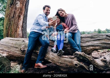 Vater nimmt Selfie über Smartphone, während er mit seiner Familie auf einem umgestürzten Baum sitzt Stockfoto