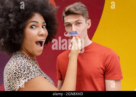 Überraschte Frau, die die Schnurrbart-Requisite über das Gesicht des Mannes hält Stockfoto