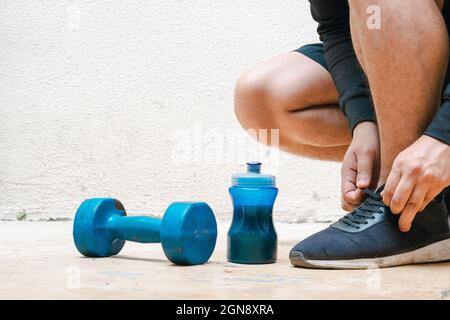 Mann bindet seine Schuhe neben Hantel und Flasche mit blauem Wasser, vor dem Training. Übung zu Hause, weißer Hintergrund. Fitnesskonzept. Gesundheit concep Stockfoto