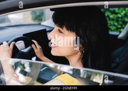 Lächelnde, reife Frau, die beim Fahren eines Elektroautos vom Fenster wegschaut Stockfoto