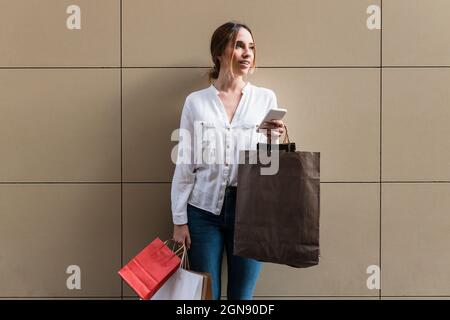 Junge Frau, die Smartphone und Papier-Einkaufstaschen vor der Wand hält Stockfoto