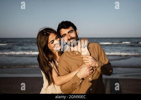 Lächelnde Frau umarmt Mann von hinten am Strand Stockfoto