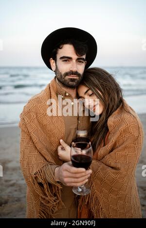 Freund in Decke gewickelt mit Freundin, die Wein am Strand hält Stockfoto