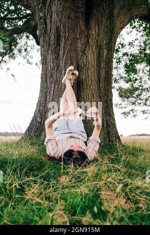 Junge Frau liest Buch, während sie unter dem Baum auf dem Gras liegt Stockfoto