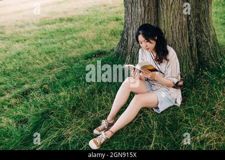 Junge Frau liest Buch, während sie unter dem Baum im Park sitzt Stockfoto