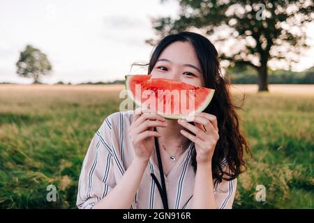 Junge Frau bedeckt das Gesicht mit einer Wassermelonenscheibe auf der Wiese Stockfoto