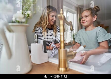 Lächelndes Mädchen, das durch das Fenster schaut, während es mit der Mutter in der Küche Geschirr abspült Stockfoto