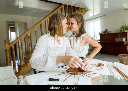 Frau lächelt Tochter an, während sie im Wohnzimmer einen Korb webt Stockfoto