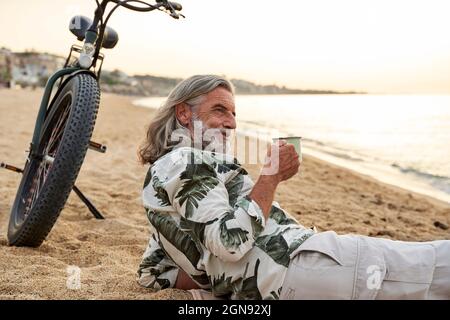 Reifer Mann hält Becher beim Entspannen am Strand Stockfoto