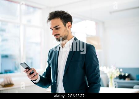 Hübscher junger Geschäftsmann, der im Büro steht und über das Smartphone im Internet surft Stockfoto