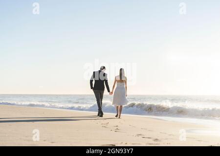 Das frisch verheiratetes Paar hielt sich die Hände, während es am Strand spazieren ging Stockfoto