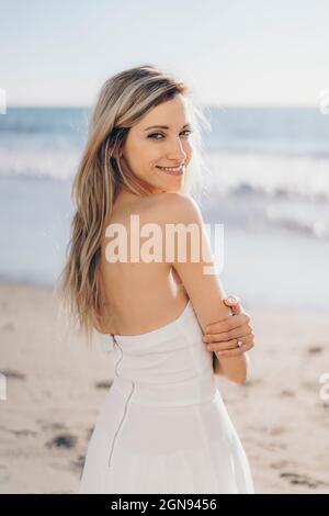 Lächelnde Braut im Hochzeitskleid, die am Strand steht Stockfoto