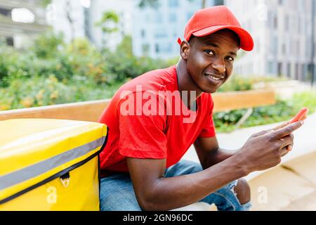 Lächelnder junger Mann, der auf der Bank saß und das Smartphone hielt Stockfoto