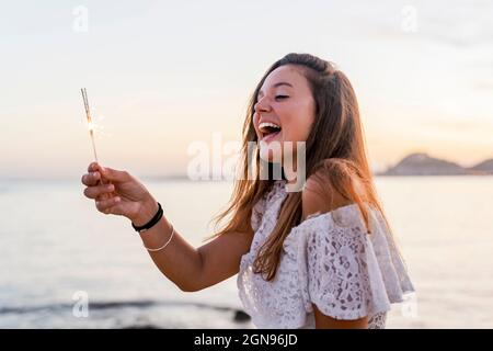 Fröhliche junge Frau, die am Strand Sparkler in der Hand hält Stockfoto