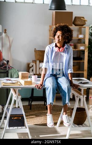 Glückliche junge Unternehmerin mit Kopfhörern, die im Studio auf dem Schreibtisch sitzen Stockfoto