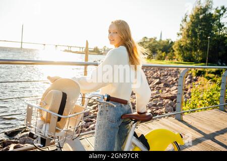 Junge blonde Frau blickt über die Schulter, während sie an sonnigen Tagen mit dem Fahrrad steht Stockfoto