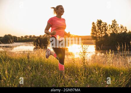 Lächelnde Frau beim Joggen auf der Wiese während des Sonnenuntergangs Stockfoto